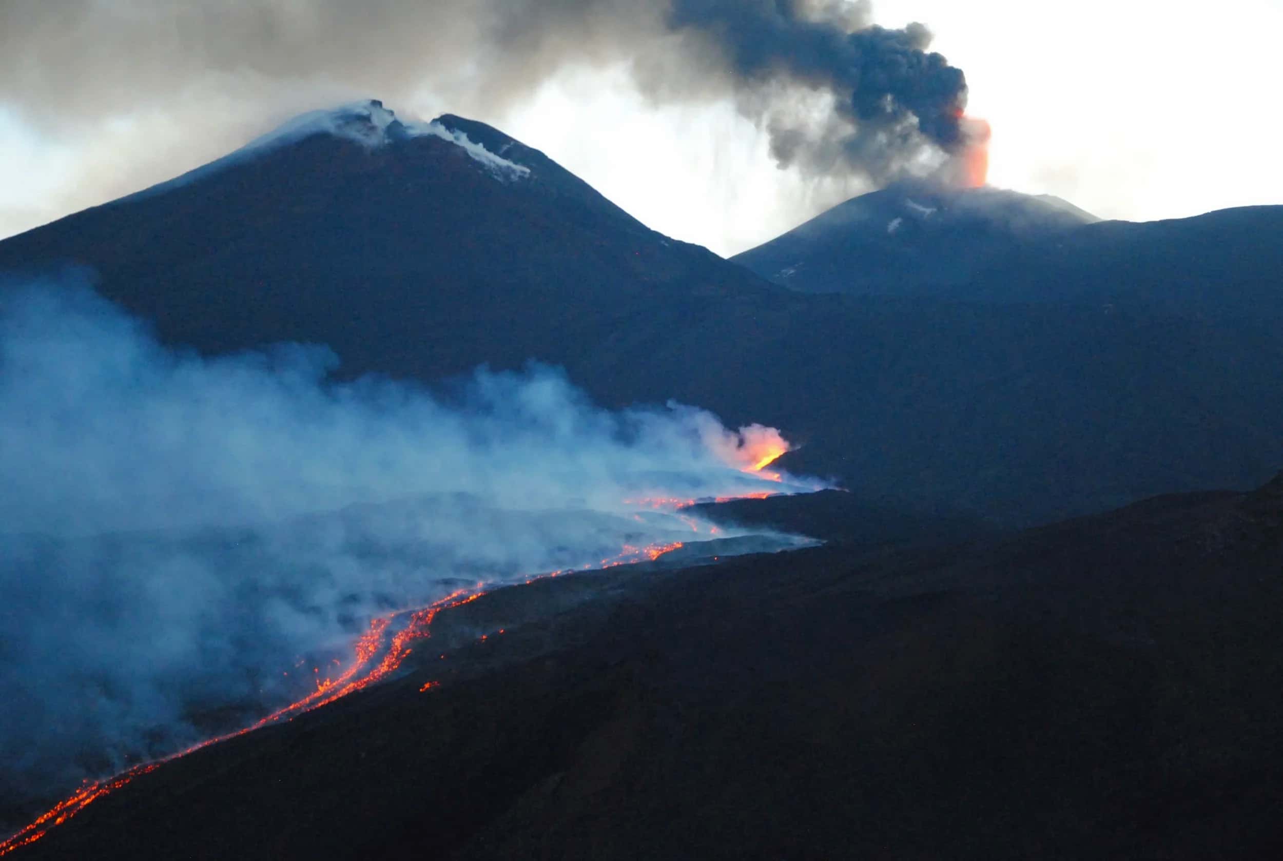 La colata lavica scorre tra i crateri al crepuscolo, tra vapori e fumo